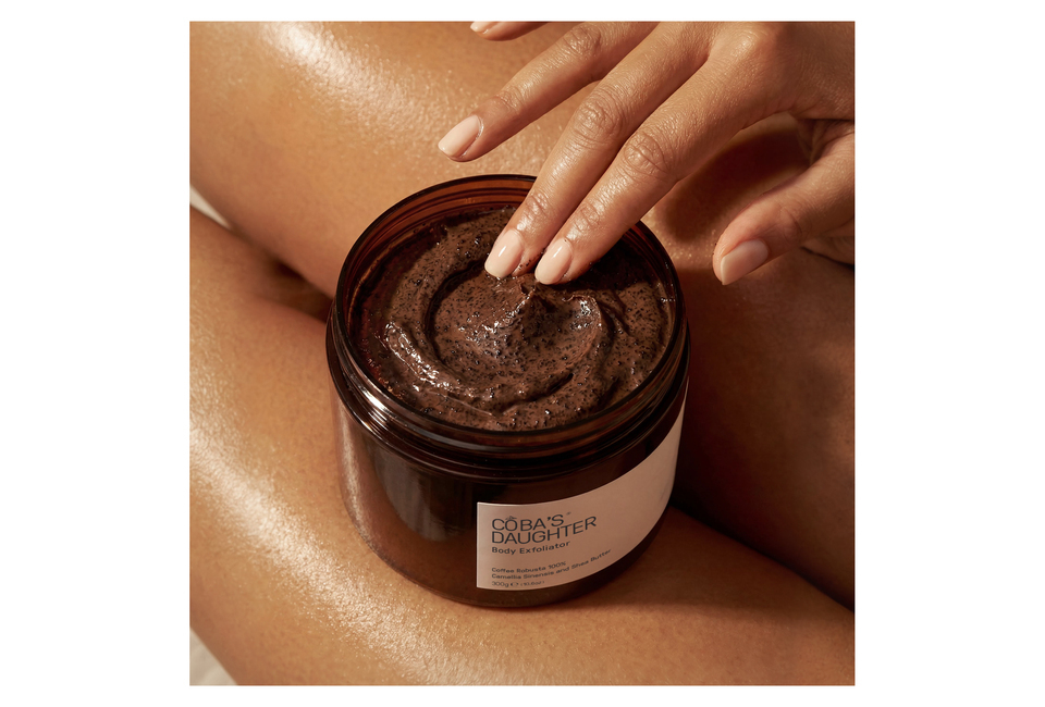 Sản phẩm CoBa’s Daughter Body Exfoliator 300gr được trưng bày với tay có độ ẩm lấy một phần bộ sản phẩm, phần da chân sáng bóng bên dưới.